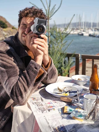 Man wearing a brown textured coat holding a vintage camera to his eye at an outdoor table by a marina.