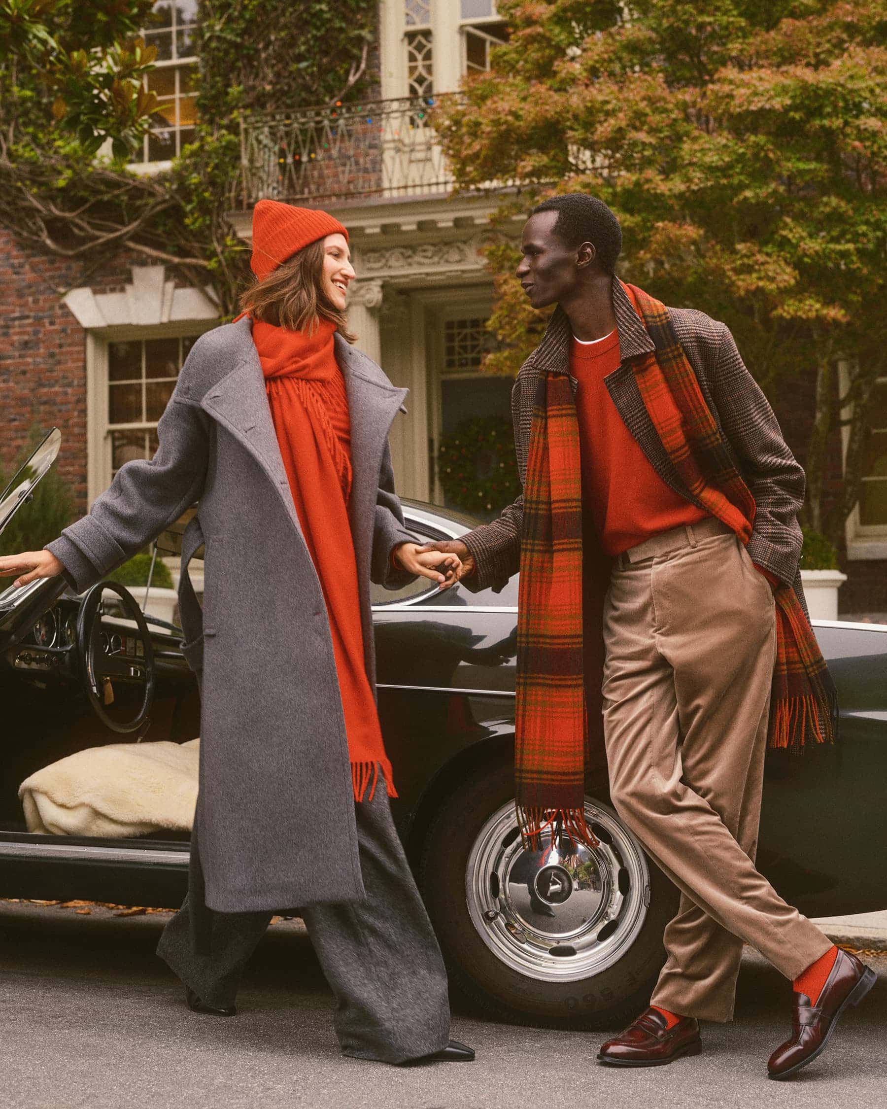 Two models wearing warm winter clothing including red scarves and coats standing next to a vintage dark car.
