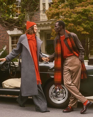 Two models wearing warm winter clothing including red scarves and coats standing next to a vintage dark car.