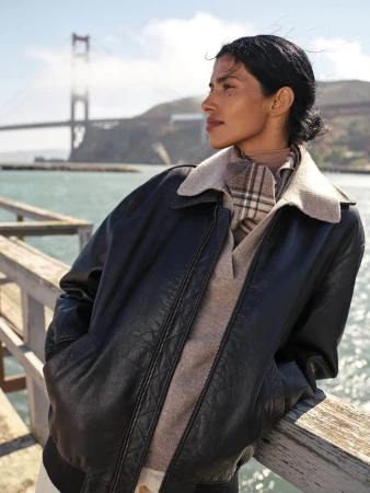 Woman in dark leather bomber jacket and plaid scarf leaning on a wooden railing with the Golden Gate Bridge visible in the background