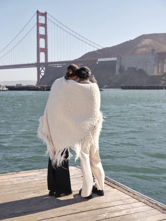 Two people wrapped in a white fringed blanket standing on a wooden pier with the Golden Gate Bridge in the background.