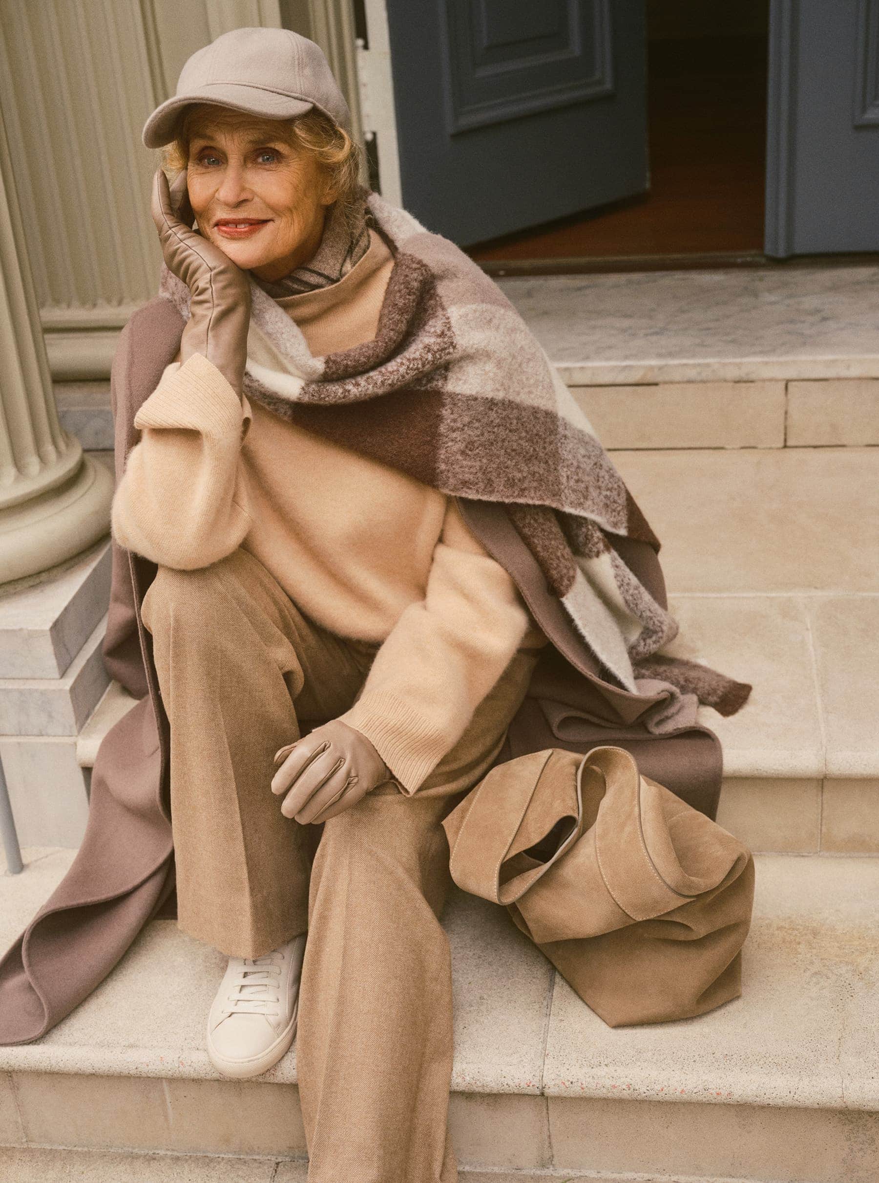 An elderly woman wearing a beige cap, trousers, and leather gloves seated on stone steps with a plaid blanket and handbag.