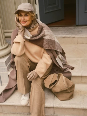An elderly woman wearing a beige cap, trousers, and leather gloves seated on stone steps with a plaid blanket and handbag.