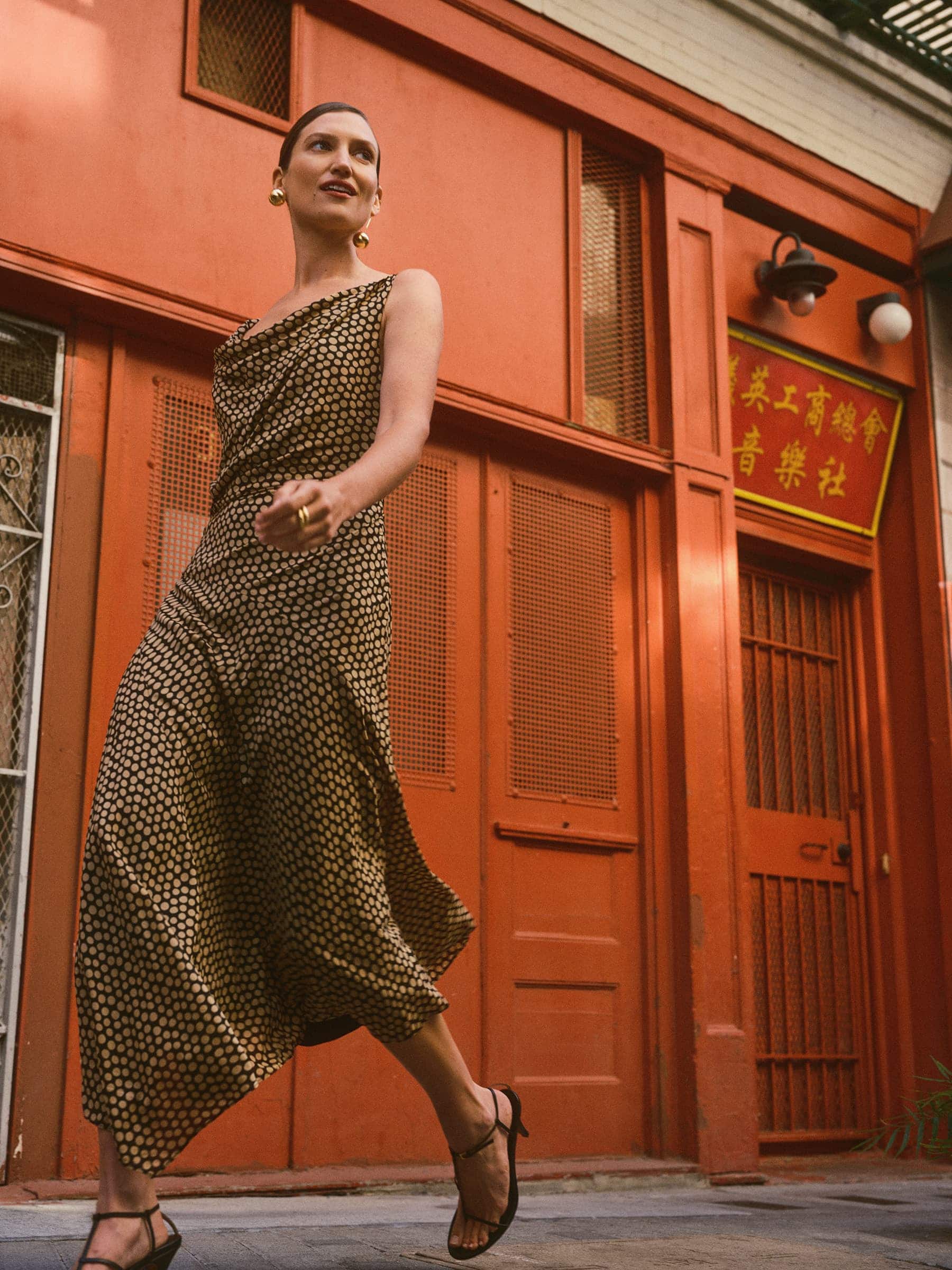 Woman in a patterned sleeveless midi dress walking past a vibrant orange building exterior.