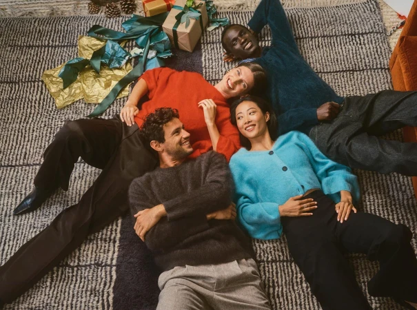 Four models lying in a circle on a patterned rug surrounded by wrapped holiday gifts.