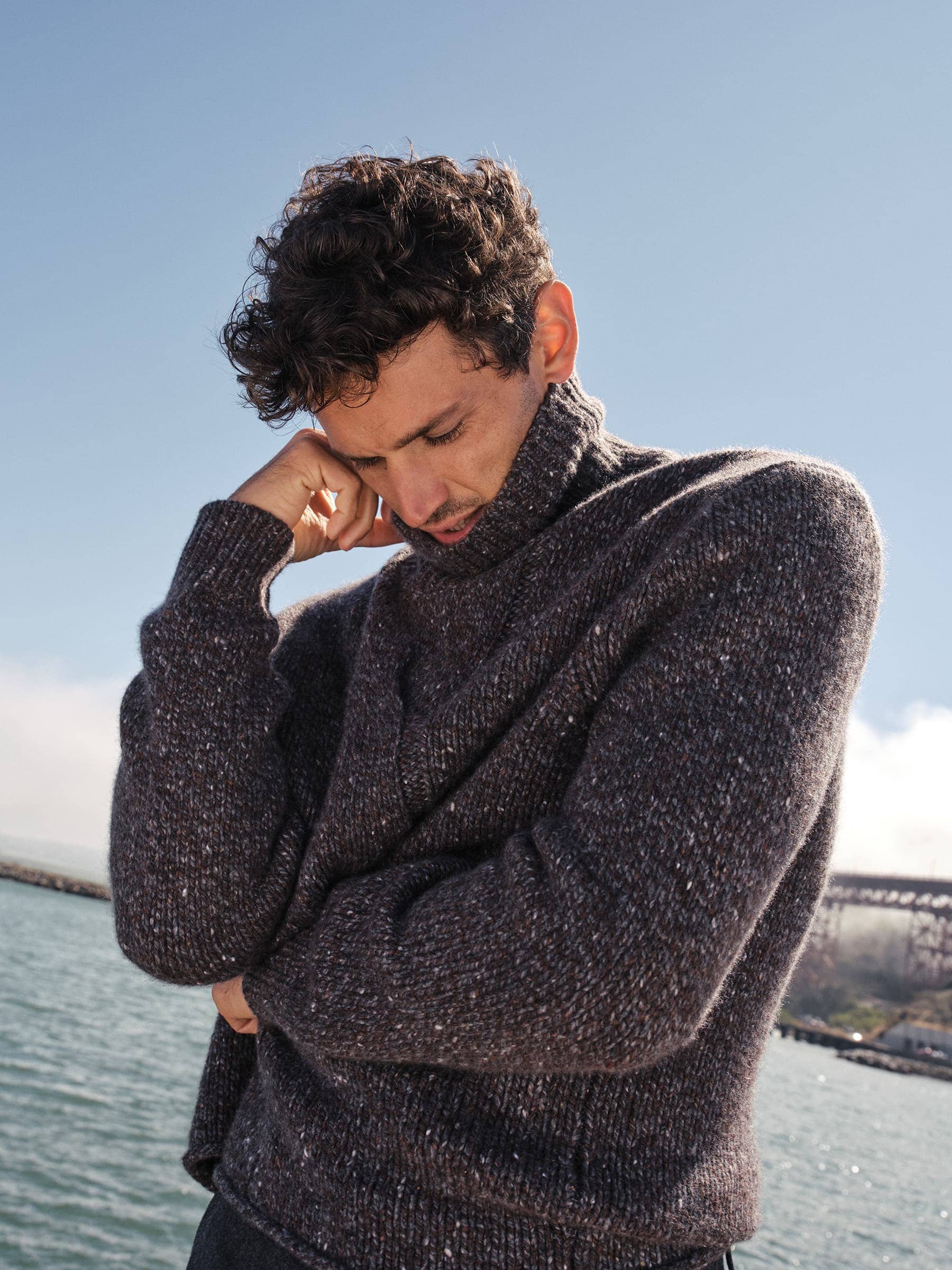 Man with curly dark hair wearing a textured dark grey turtleneck sweater outdoors near water under a clear blue sky