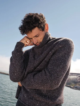 Man with curly dark hair wearing a textured dark grey turtleneck sweater outdoors near water under a clear blue sky