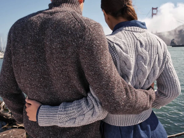 Man and woman embracing with arms around each other, wearing knitted sweaters outdoors by the water with a bridge in the background.