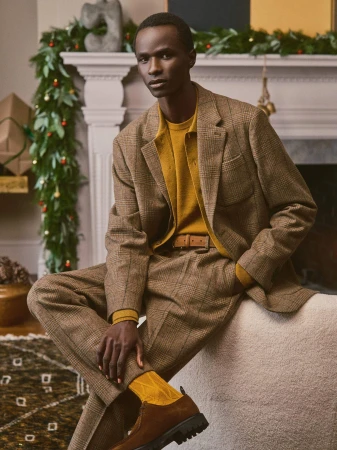 Man wearing a brown plaid suit and mustard knit sweater sitting indoors by a fireplace with holiday garland.