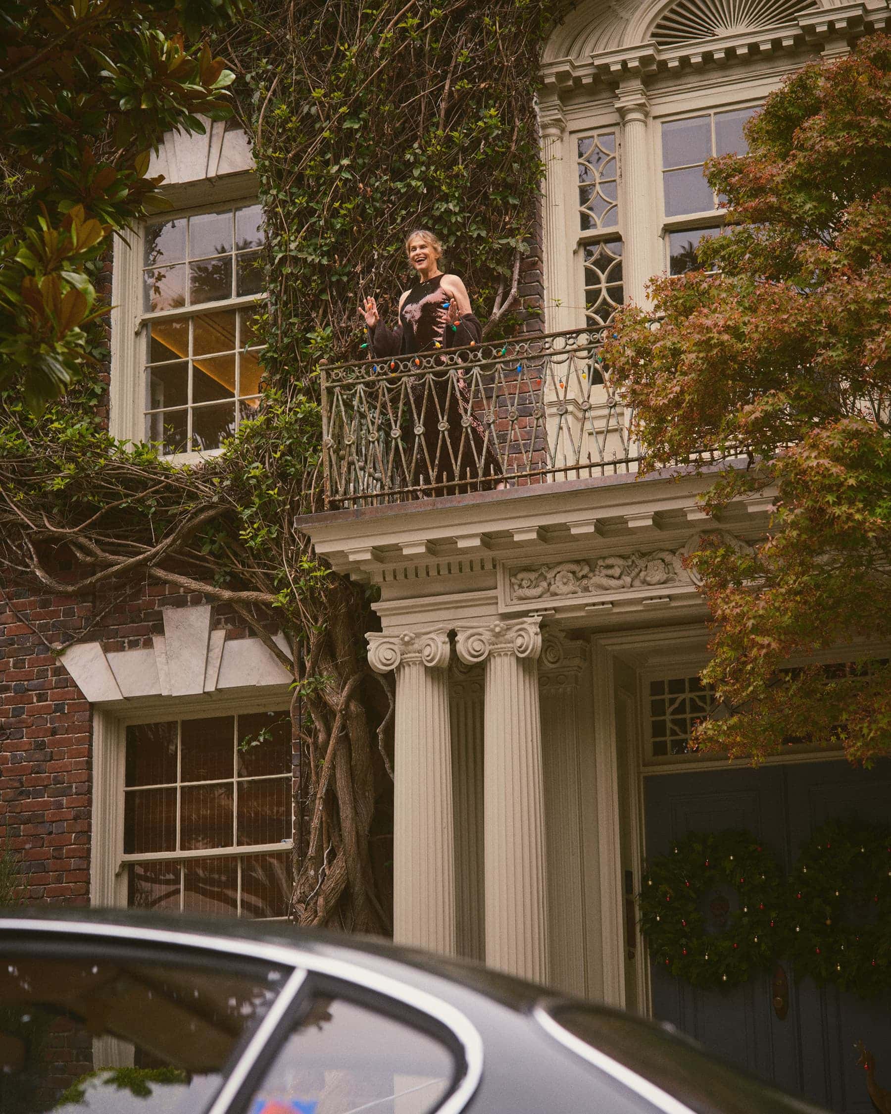 Woman standing on a classical building balcony framed by lush vines above a dark car roof.