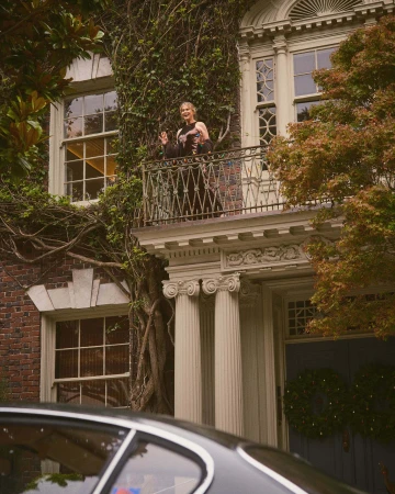 Woman standing on a classical building balcony framed by lush vines above a dark car roof.