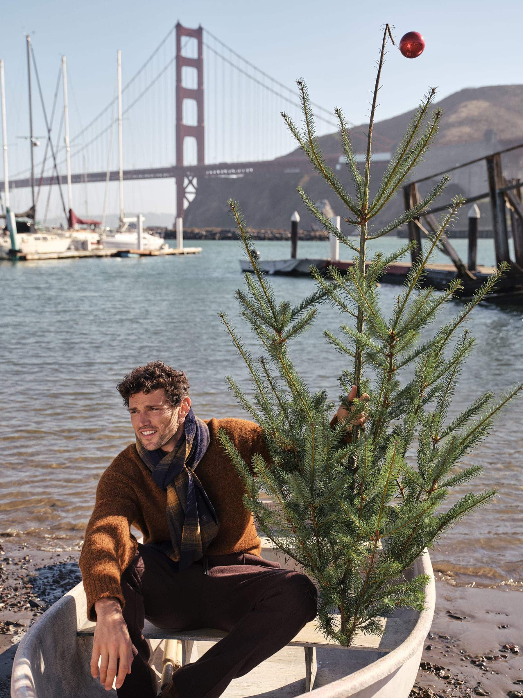 Man sitting in a small boat holding a sparse pine tree decorated with a single red ornament, with the Golden Gate Bridge in the background.
