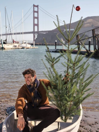 Man sitting in a small boat holding a sparse pine tree decorated with a single red ornament, with the Golden Gate Bridge in the background.
