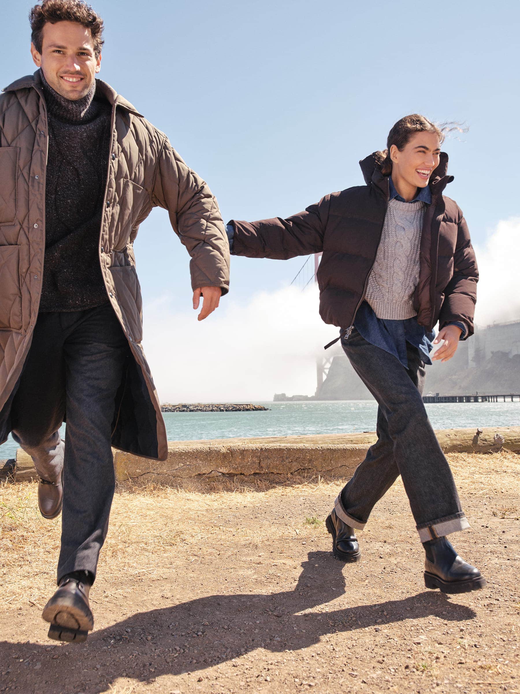Two smiling models running outdoors near a coastal waterfront wearing winter coats and dark trousers for Banana Republic.