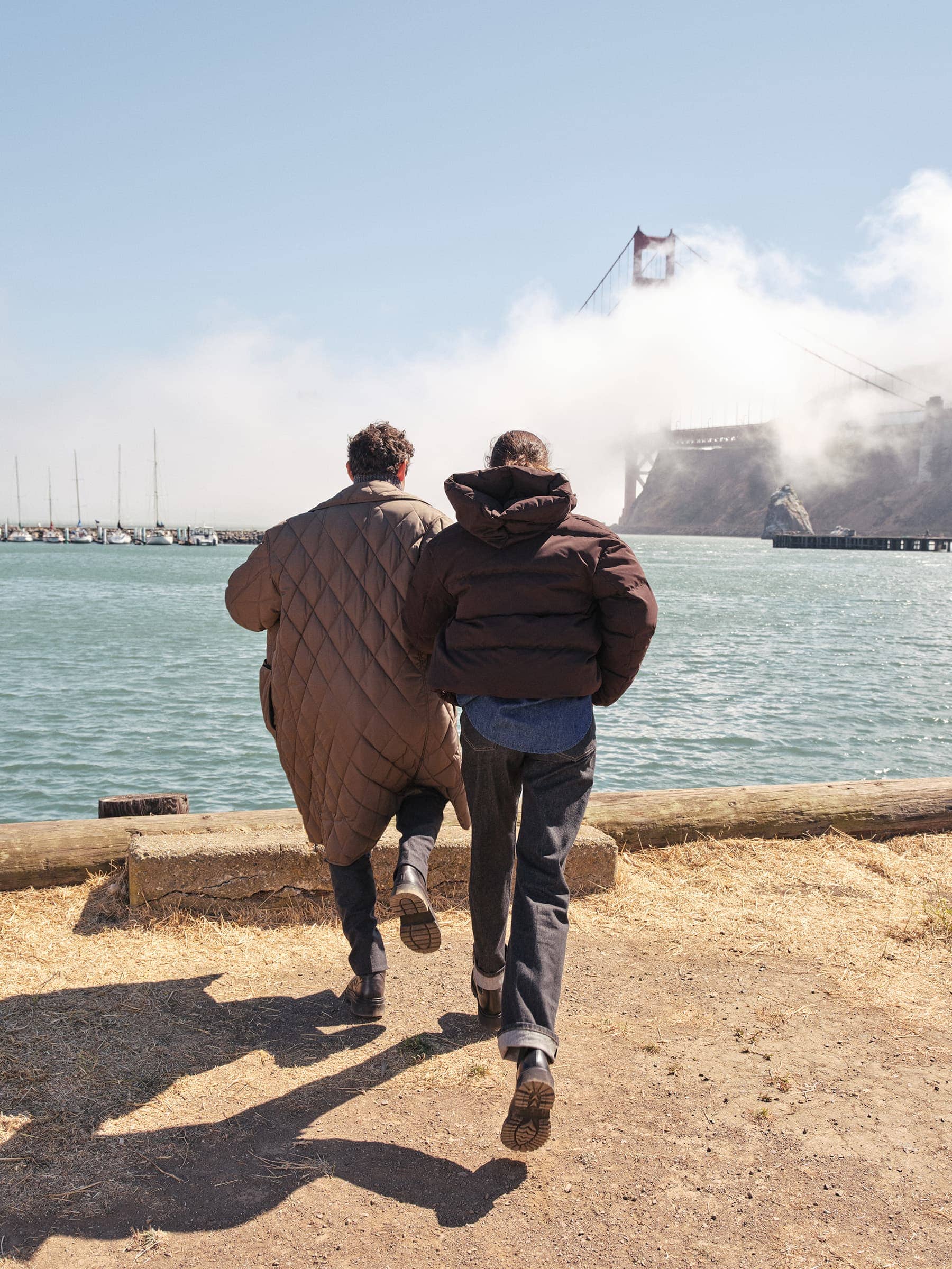 Two people wearing winter coats walking towards a coastal view with a suspension bridge partially obscured by fog.