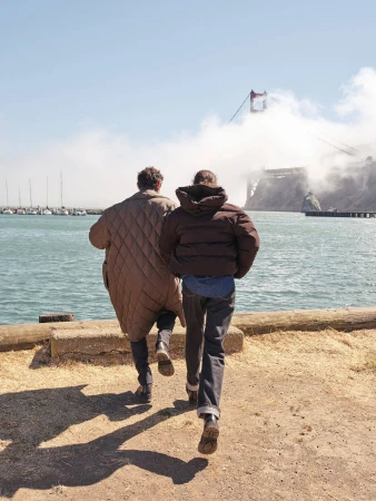 Two people wearing winter coats walking towards a coastal view with a suspension bridge partially obscured by fog.
