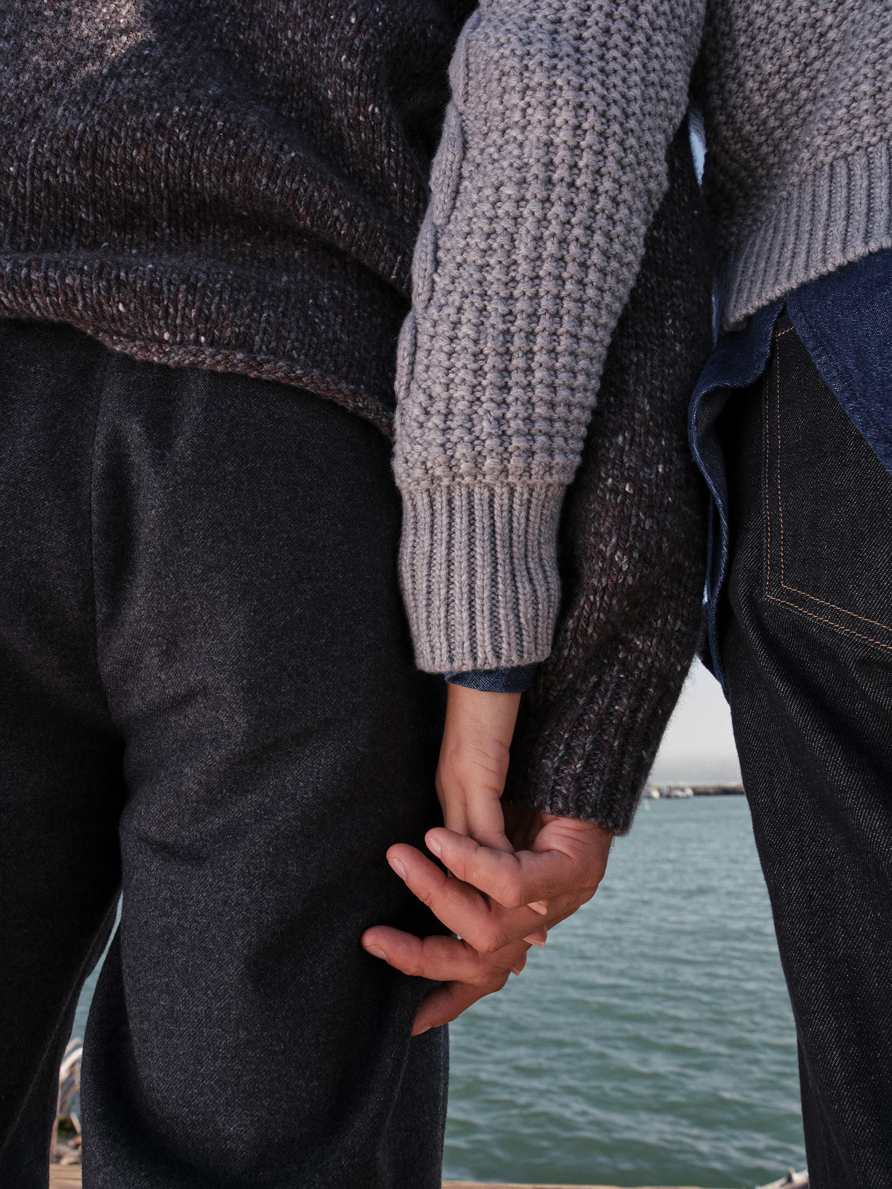 Close-up of two people holding hands wearing textured knit sweaters against a coastal water background