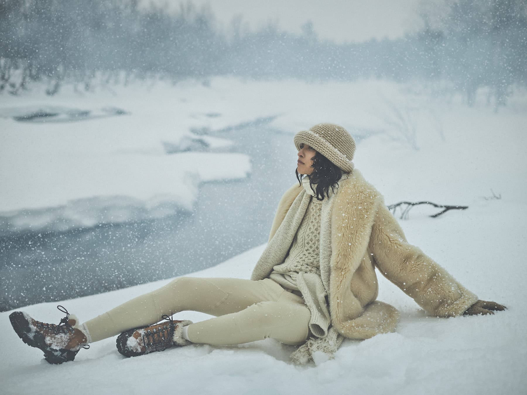 Model wearing a cream shearling coat and knit hat sitting in a snowy landscape by a frozen river