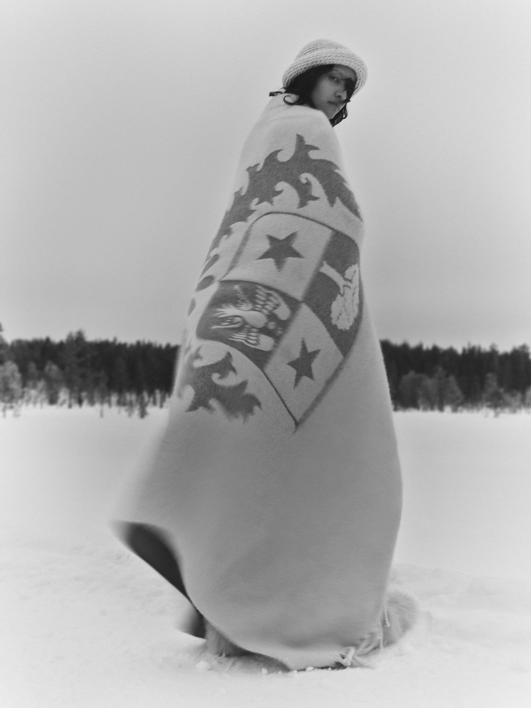 Model wrapped in a patterned blanket wearing a knit beanie standing in a snowy landscape under an overcast sky.