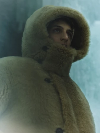 Close-up portrait of a man wearing a plush beige hooded winter coat against an icy background.