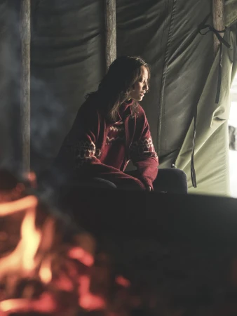 Woman in a burgundy patterned turtleneck sweater seated inside a tent near a glowing fire for Loro Piana