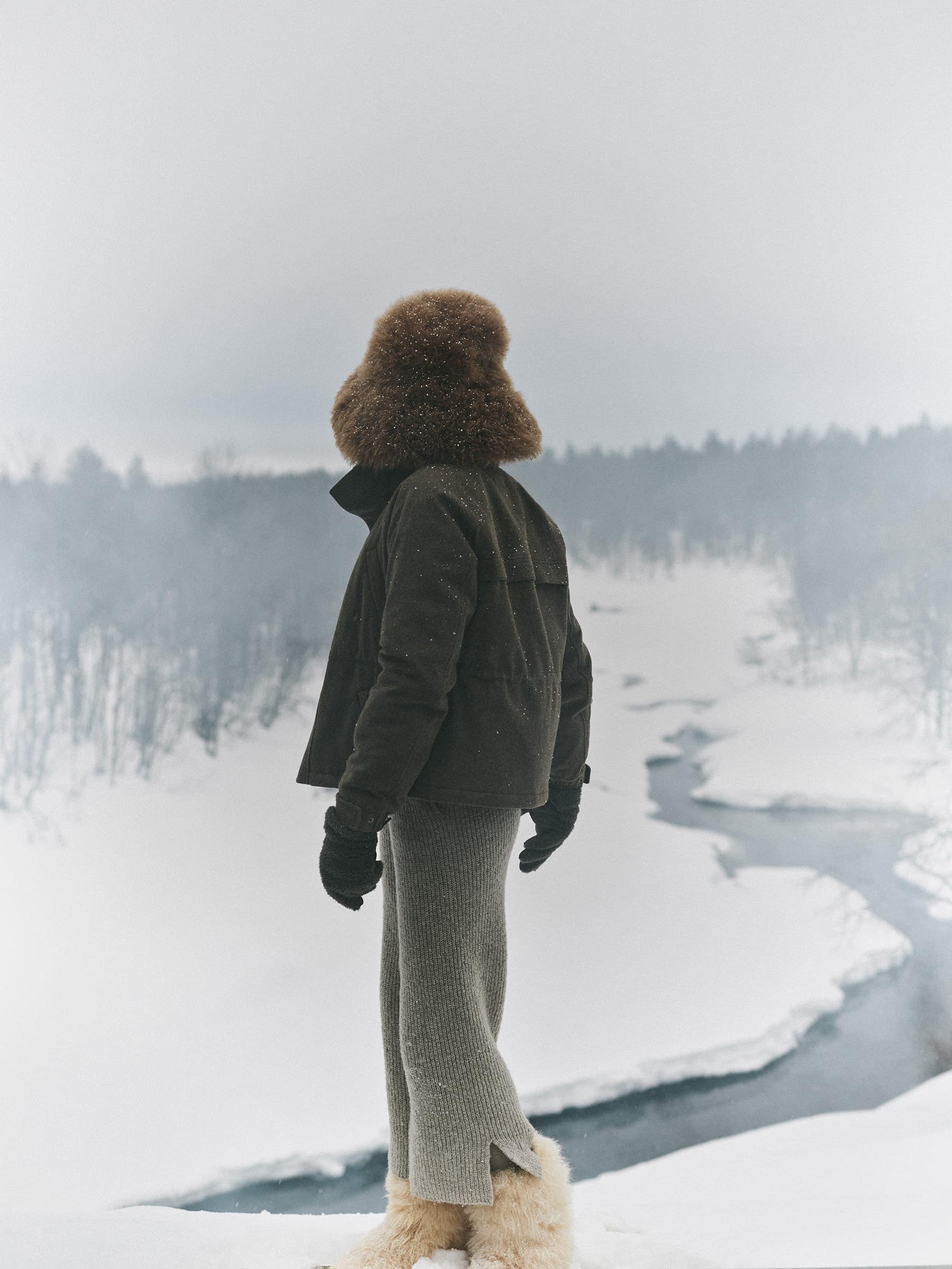 Figure standing in a snowy landscape wearing a dark jacket, fur hat, and light skirt overlooking a river.