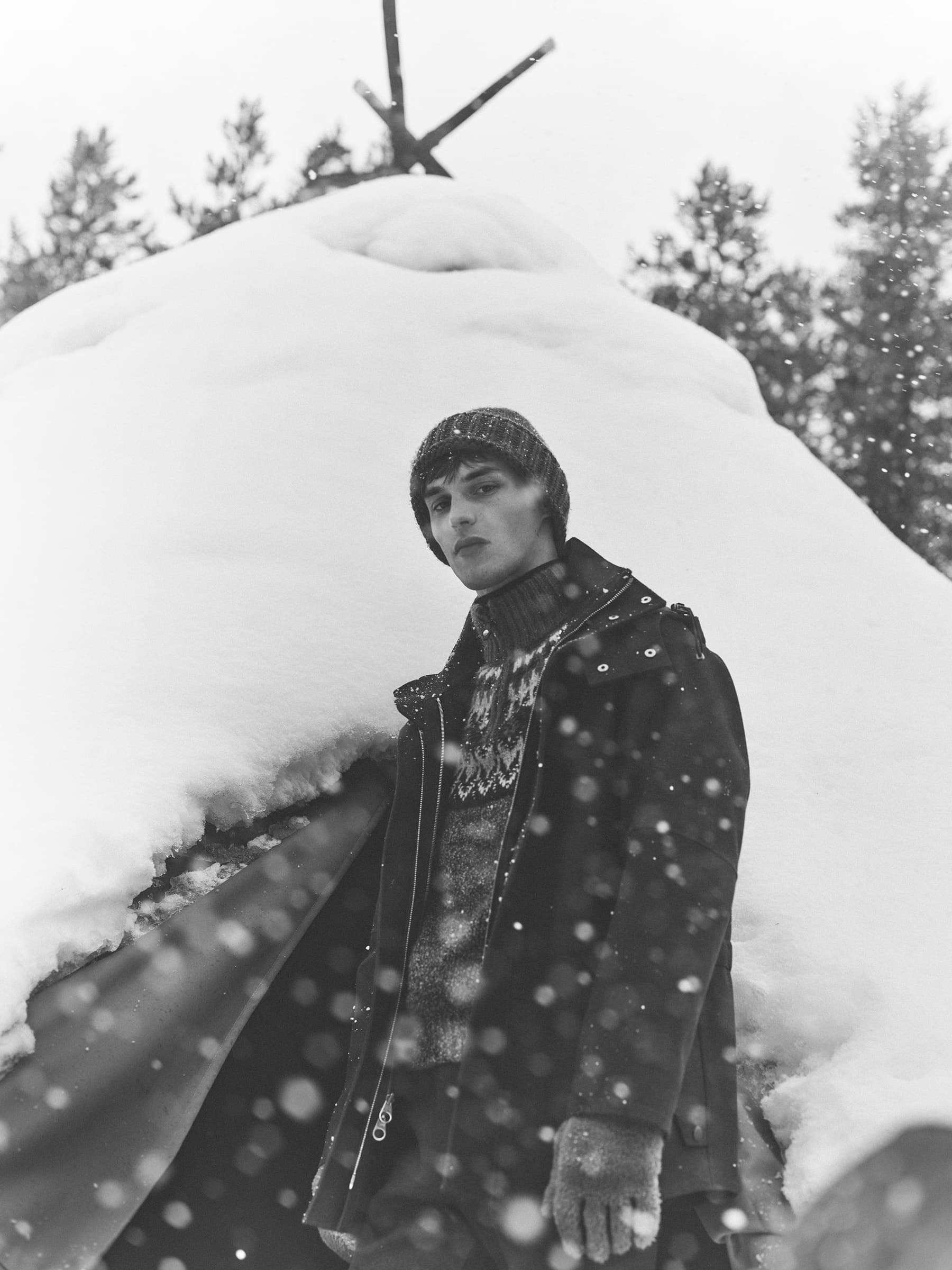 Man wearing a textured beanie and dark winter jacket standing outdoors in front of a snow-covered shelter