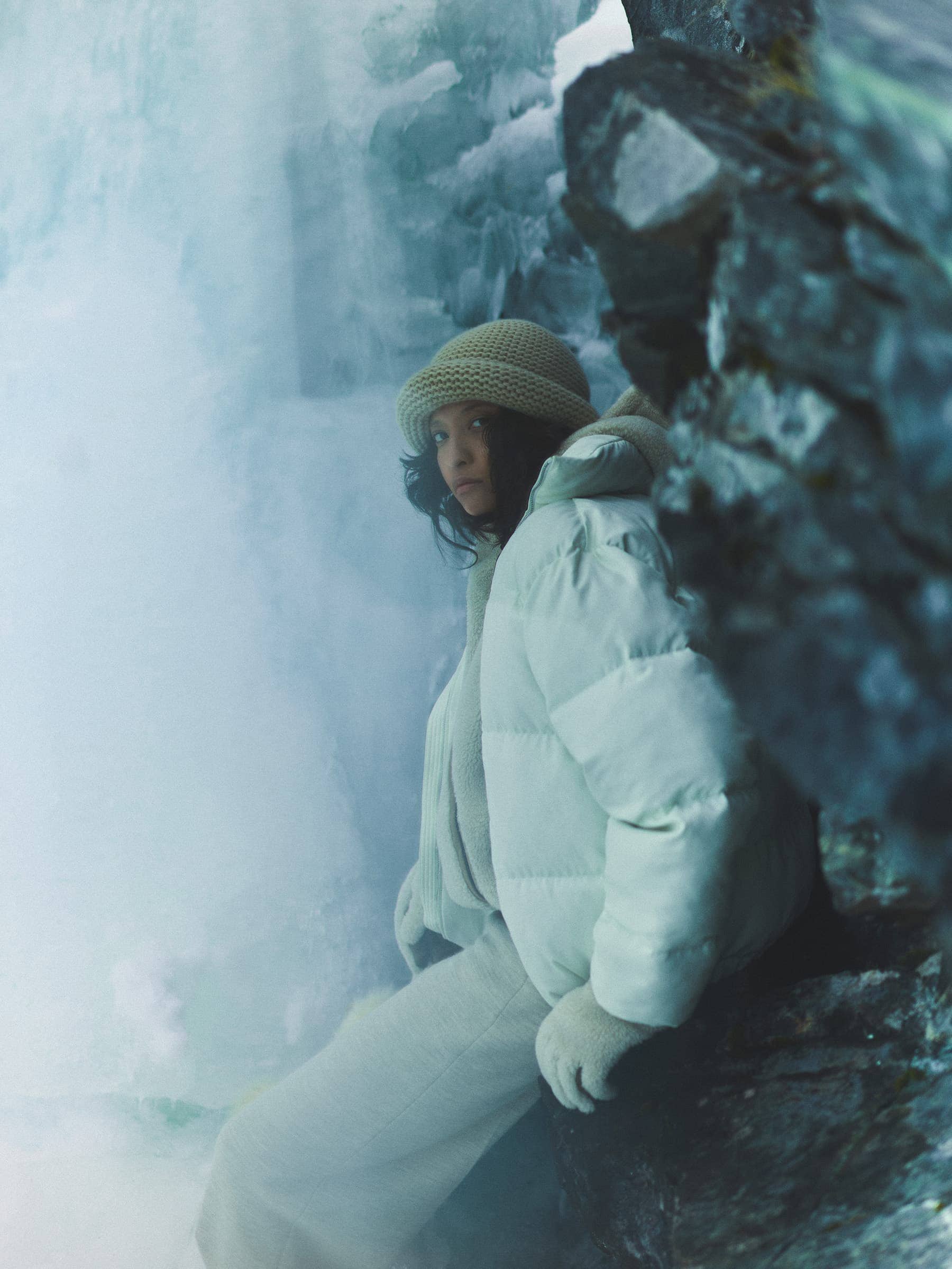 Model wearing a white puffer jacket and knit hat posing near icy rocks and a frozen waterfall.