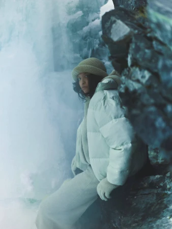 Model wearing a white puffer jacket and knit hat posing near icy rocks and a frozen waterfall.