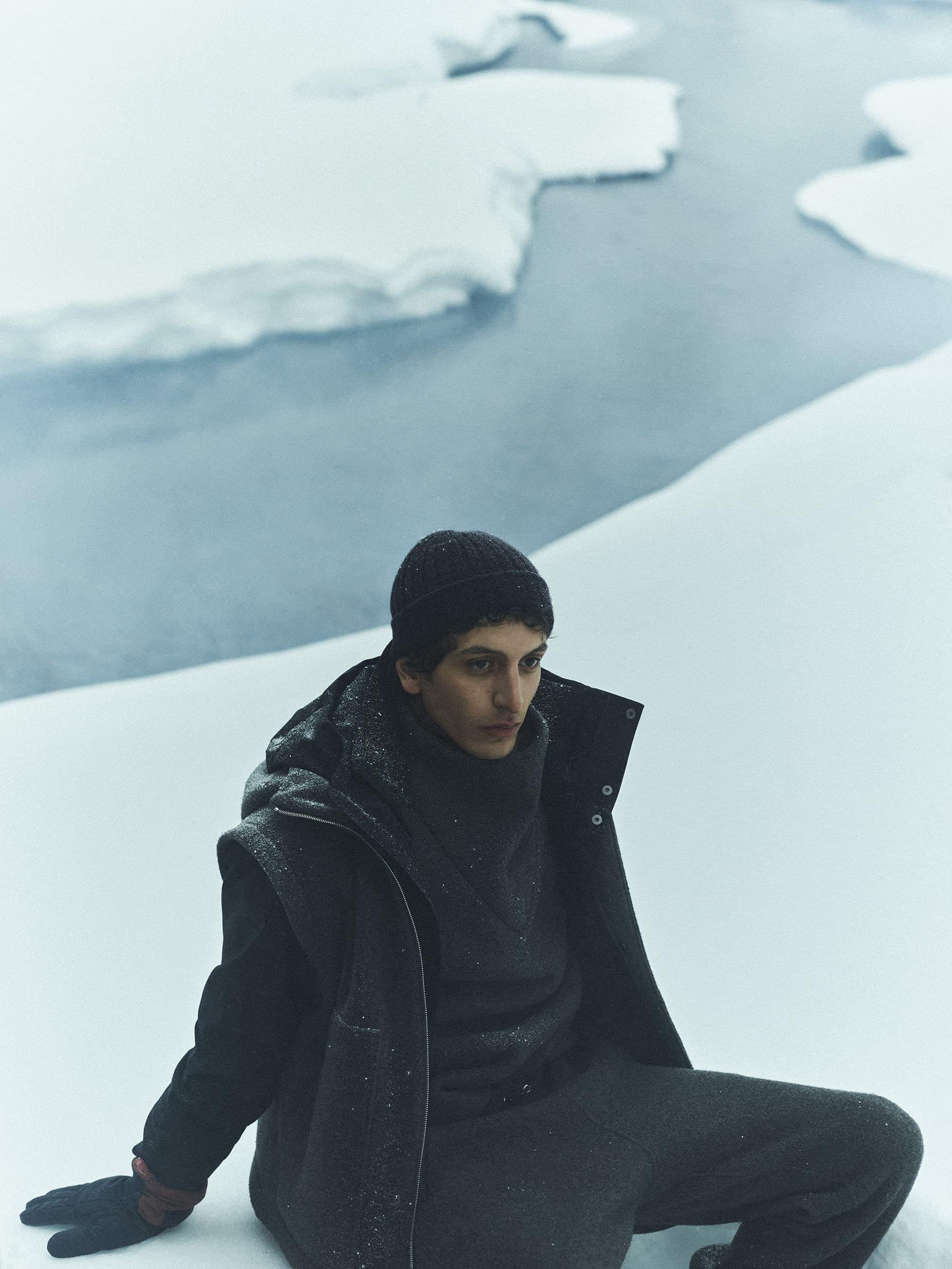 A man wearing a dark hooded vest, dark beanie, and gloves sitting on a snowy terrain overlooking water.