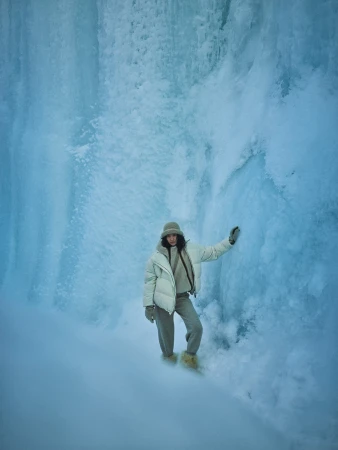 Woman in winter outerwear posing against a massive vertical wall of blue ice and snow formations