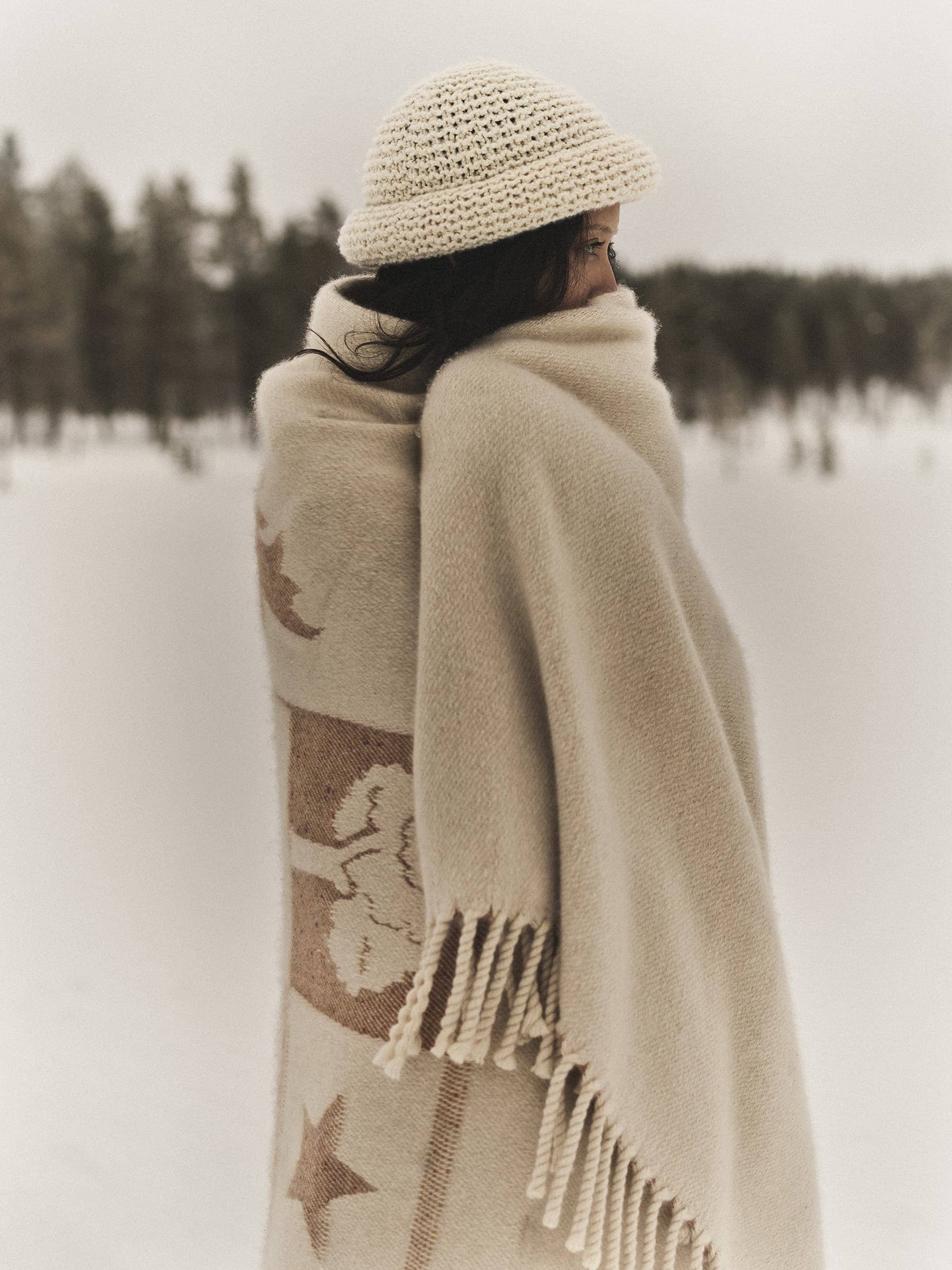 Model wrapped in a patterned cream wool blanket wearing a knitted beanie in a snowy landscape for Loro Piana.
