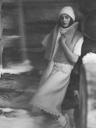 Woman in knit hat and winter scarf posing near a rustic wooden cabin for Loro Piana