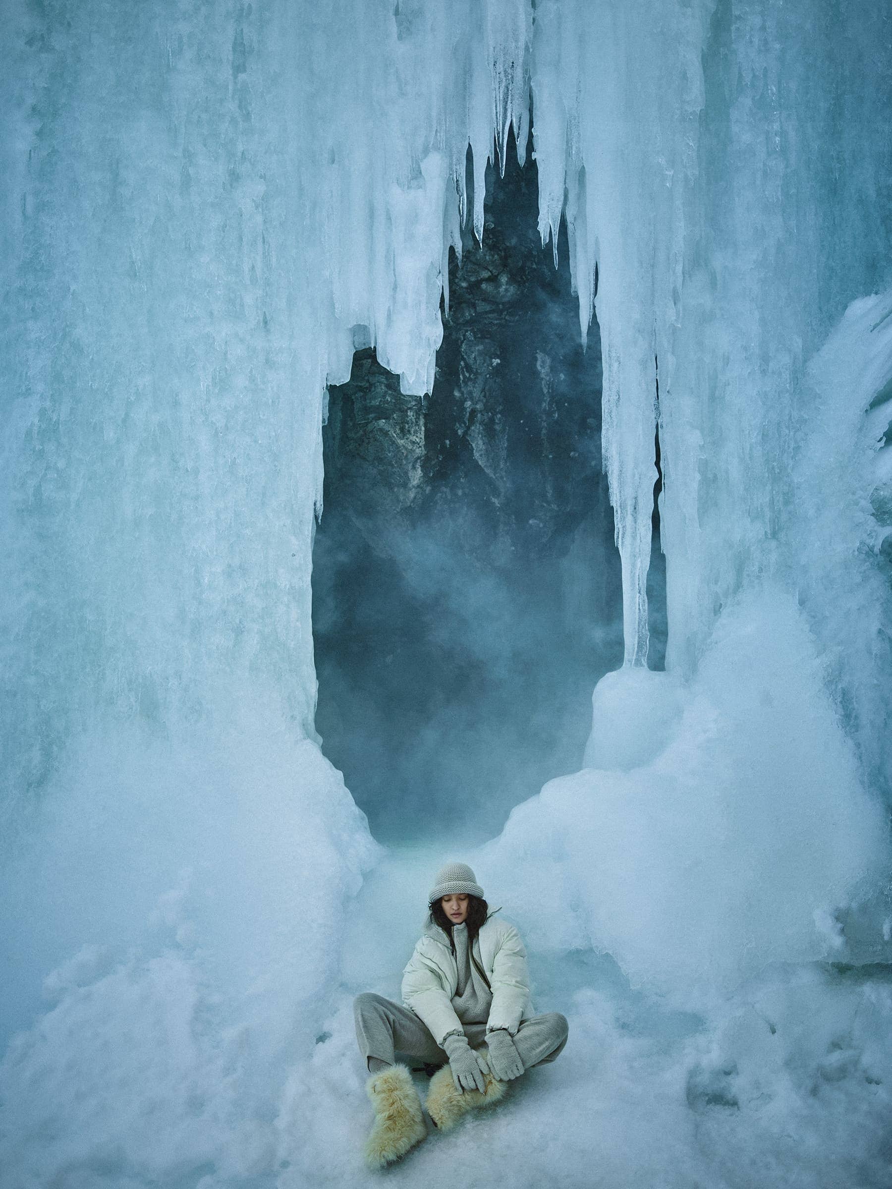 Model seated cross-legged in winter apparel beneath hanging icicles in a glacier cave