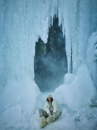 Model seated cross-legged in winter apparel beneath hanging icicles in a glacier cave