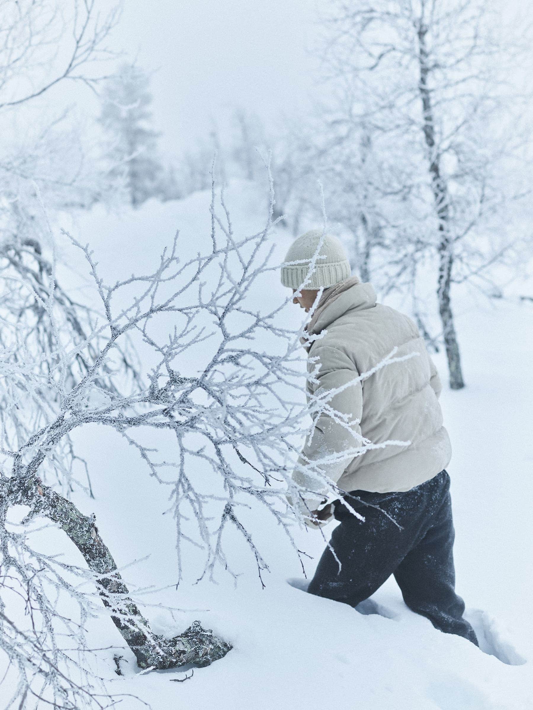 Person wearing a cream puffer jacket and beanie wading through deep snow in a frosty winter forest.
