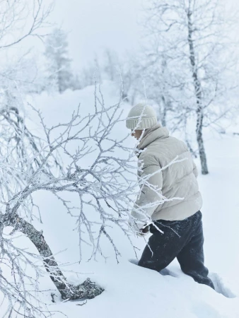 Person wearing a cream puffer jacket and beanie wading through deep snow in a frosty winter forest.