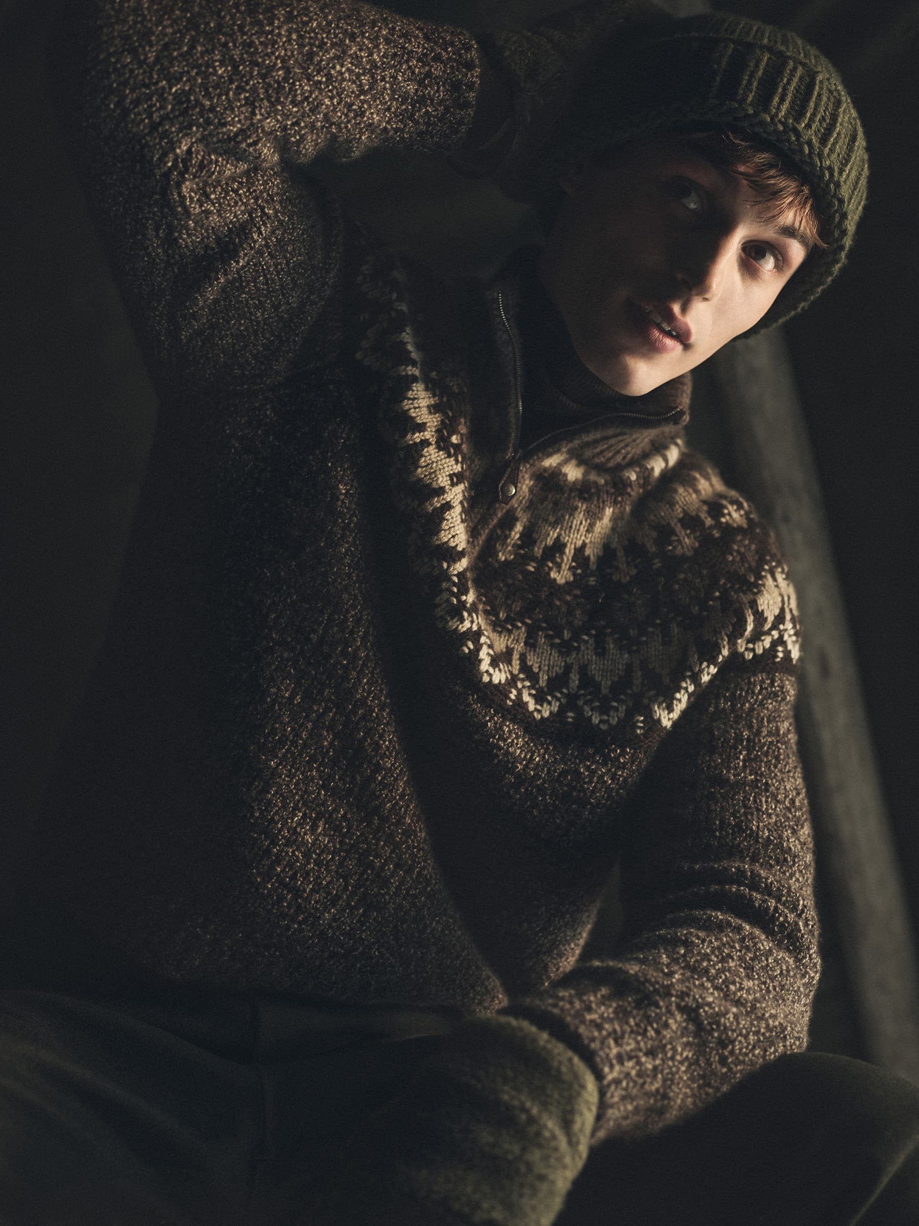 Man wearing a patterned brown knit sweater and green beanie in a dimly lit indoor setting.