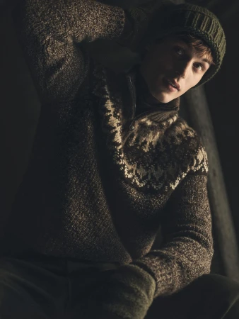 Man wearing a patterned brown knit sweater and green beanie in a dimly lit indoor setting.