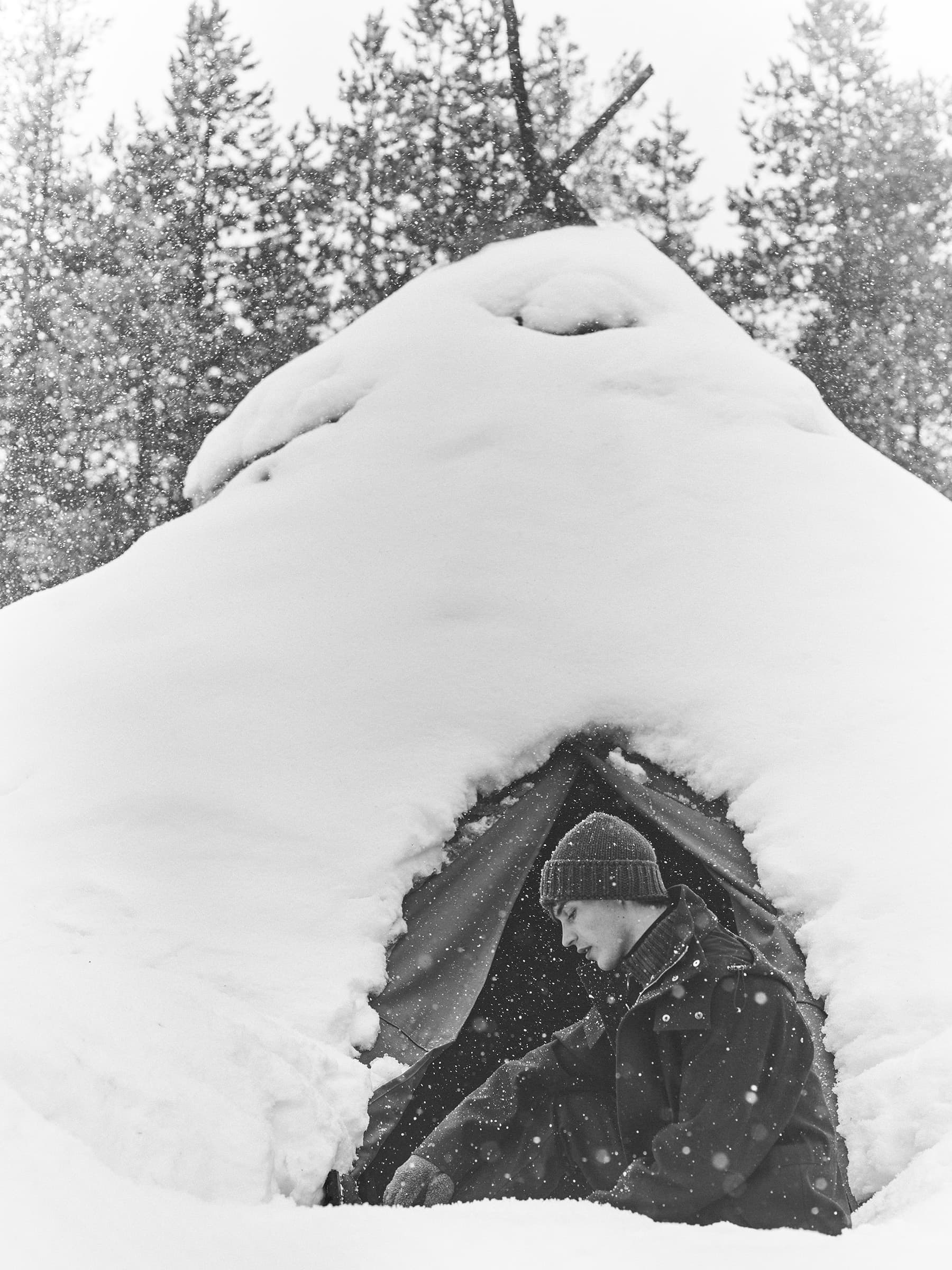 Monochrome image of a man in a beanie and heavy coat seated inside a snow-covered tent beneath pine trees.