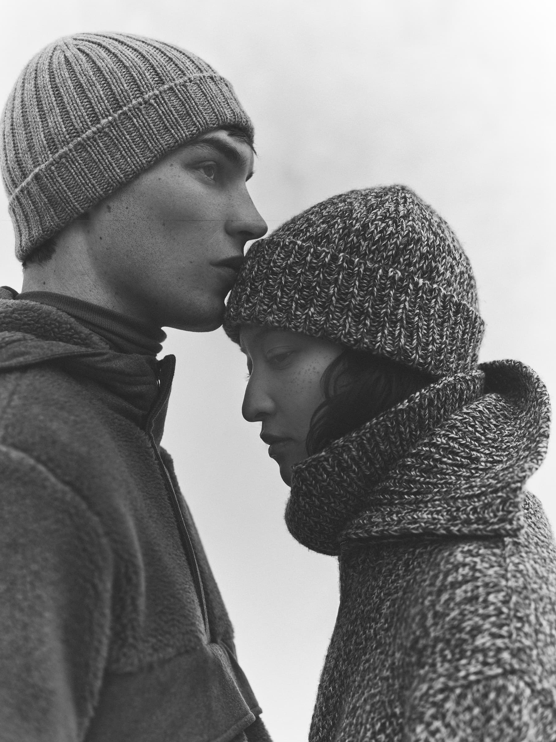 Man and woman wearing knit beanies and winter outerwear with heads touching outdoors against an overcast sky