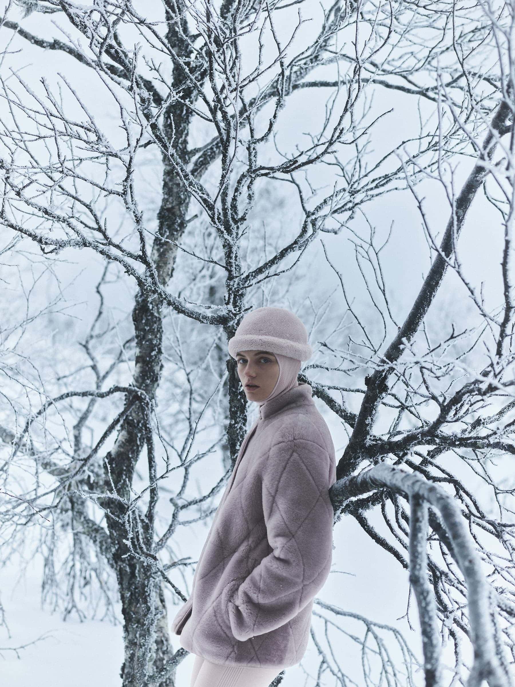 Model wearing a pink quilted jacket and matching hat leaning against a snow-covered tree in a winter forest.