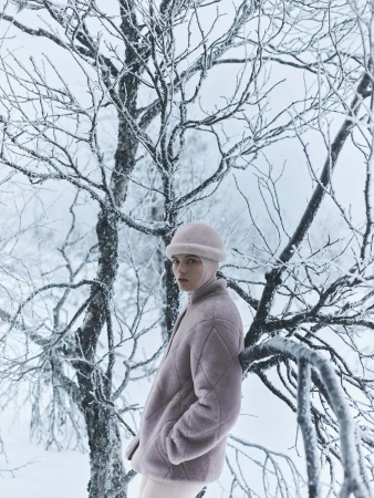 Model wearing a pink quilted jacket and matching hat leaning against a snow-covered tree in a winter forest.