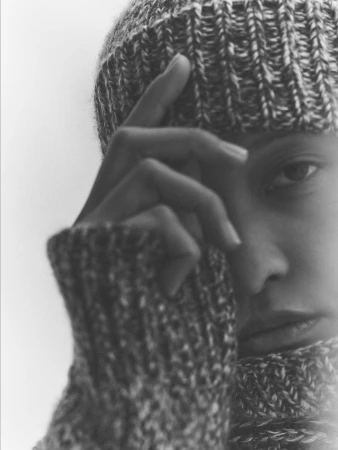 Close-up portrait of a woman wearing a heavy knit beanie and ribbed sweater against a plain background