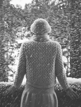Monochrome rear view of a person wearing a knit sweater and beanie outdoors amidst falling snow.