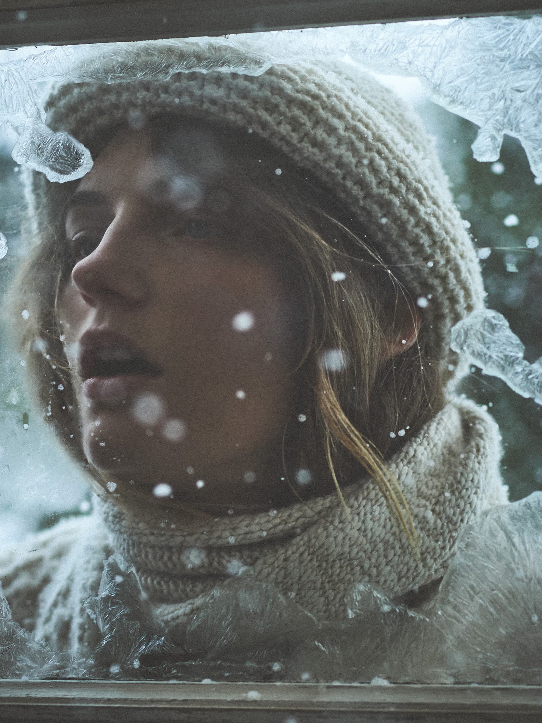 Close-up portrait of a woman wearing a cream knit cap and matching scarf seen through a frosty window pane