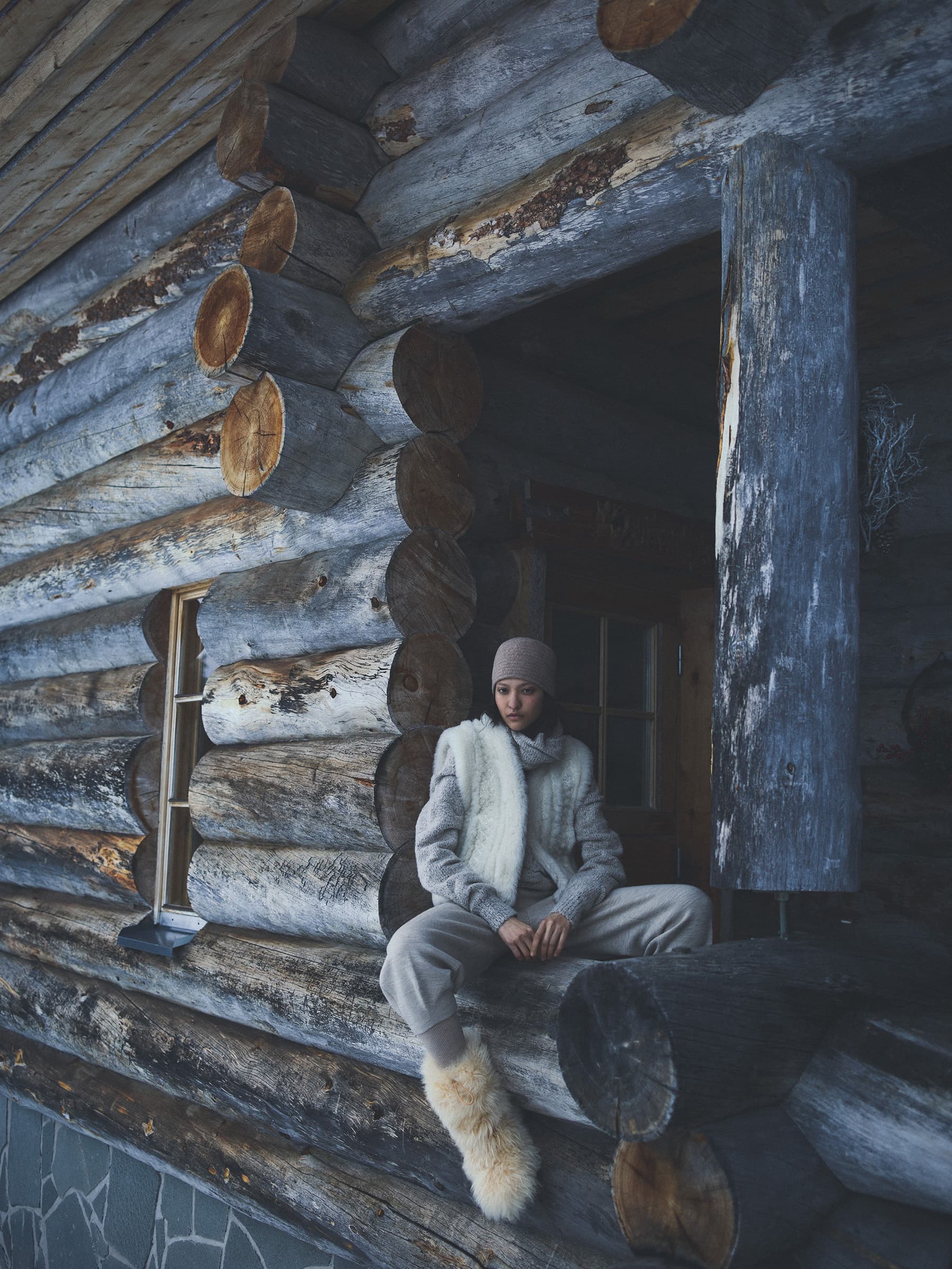 Woman in cream winter knitwear seated on the rustic log cabin porch for Loro Piana campaign