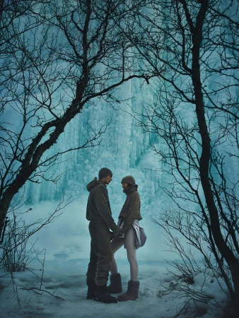 Two models standing outdoors framed by bare winter trees against a massive frozen blue ice waterfall wearing warm apparel