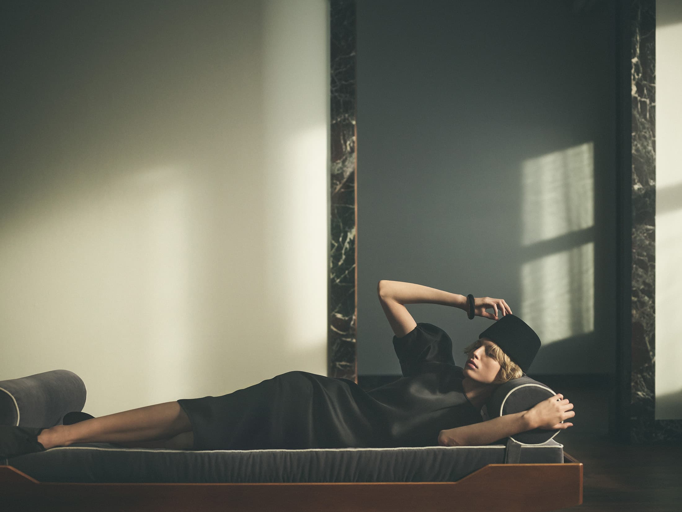 Model lying on a daybed wearing a dark dress and pillbox hat in a sunlit interior space