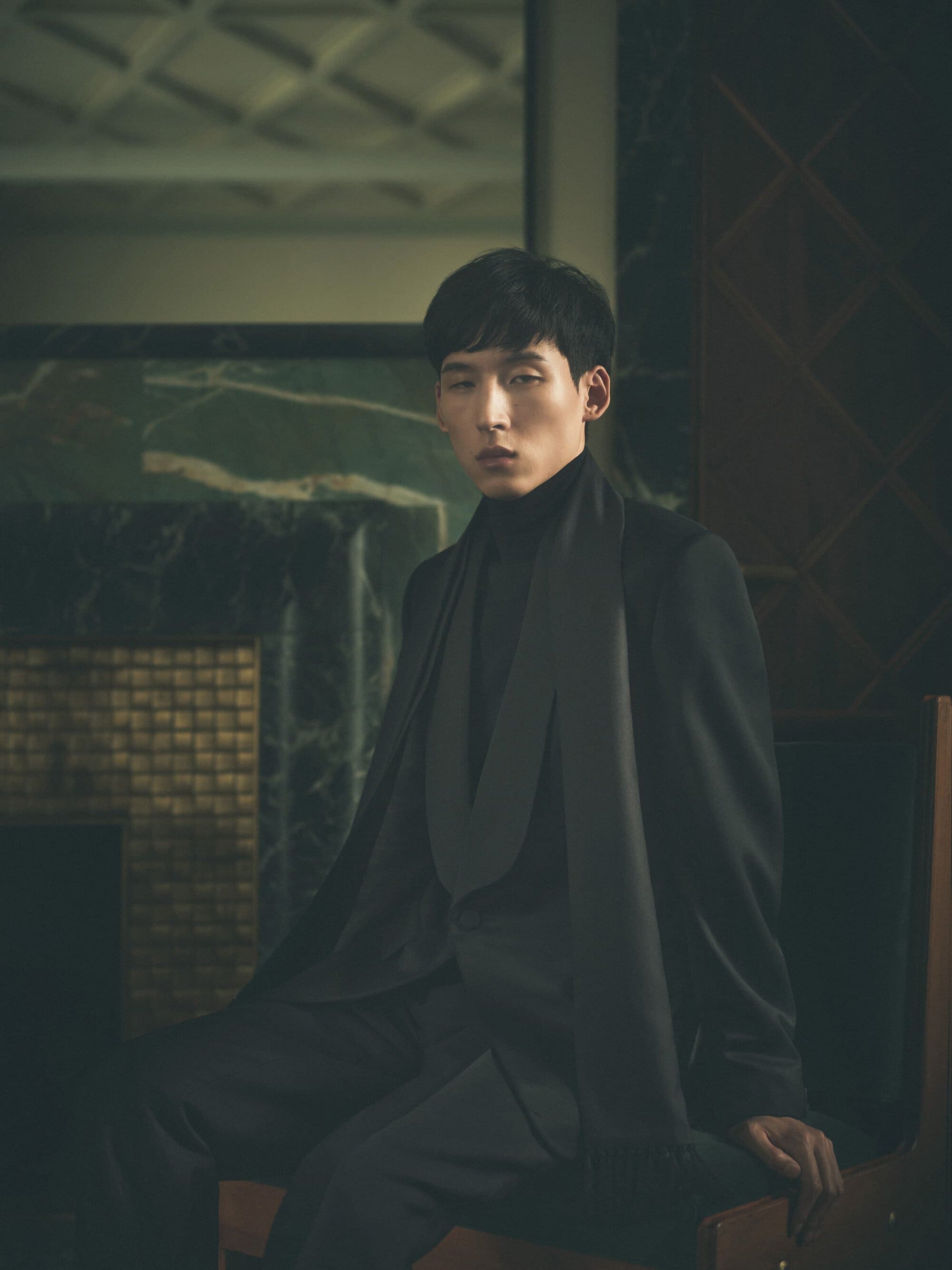 Man wearing a dark suit jacket and turtleneck seated indoors against a moody interior background.
